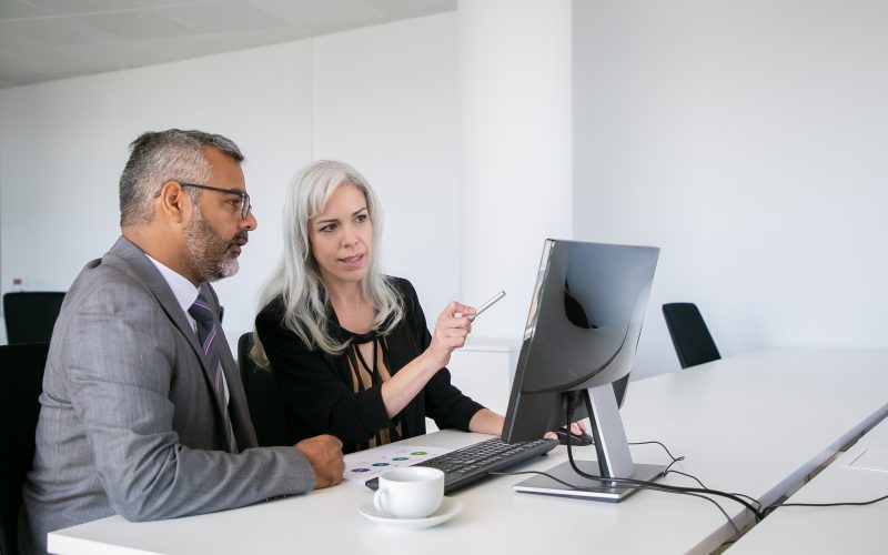 Focused business colleagues watching content on computer, pointing at display and talking while sitting at desk with paper chart. Business communication concept